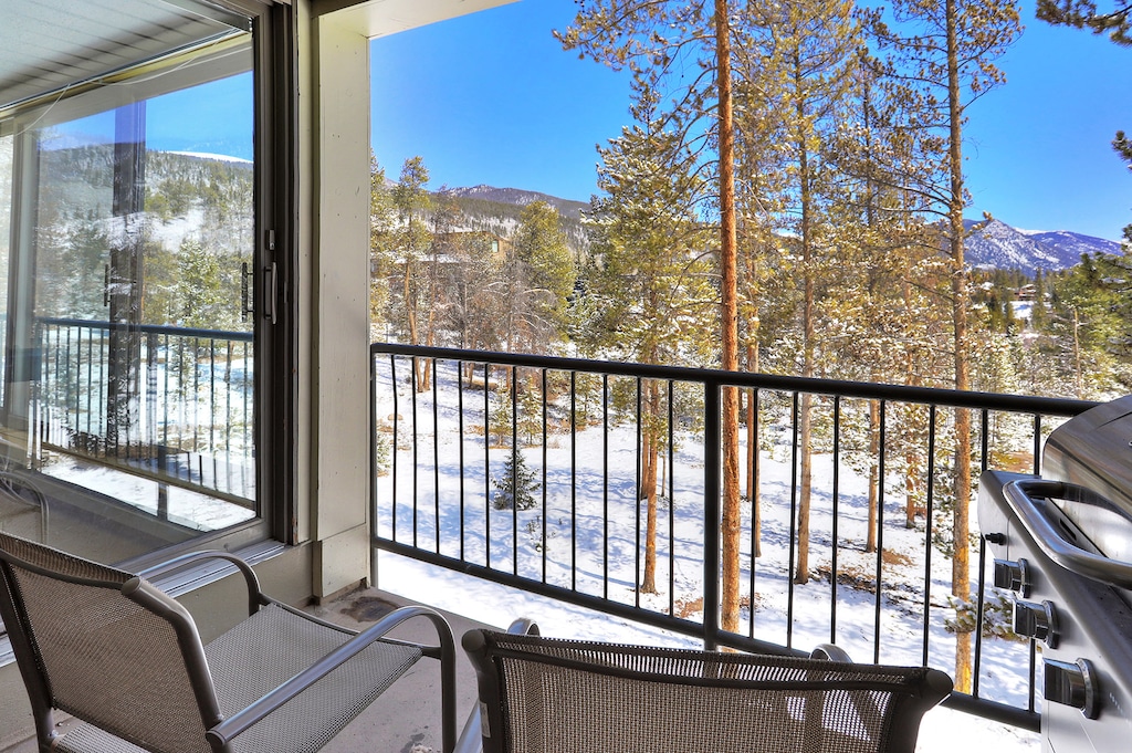 A balcony with two chairs, a barbecue grill, and a scenic view of snow-covered trees and distant mountains on a clear day.