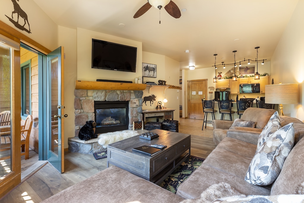 Living room with a stone fireplace, mounted TV, and sectional sofa. Wooden coffee table in center. Open concept connects to the kitchen with bar stools. Balcony door open, showing a patio.