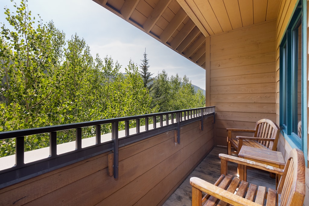 A wooden deck with chairs overlooks green trees and a forested landscape. The deck has a wooden ceiling and railing.