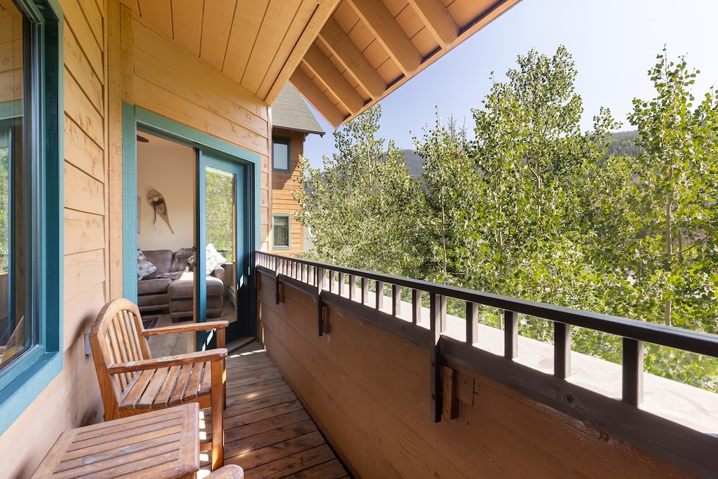 A wooden balcony with two chairs overlooks green trees. A partially open door leads inside to a living room with a couch.