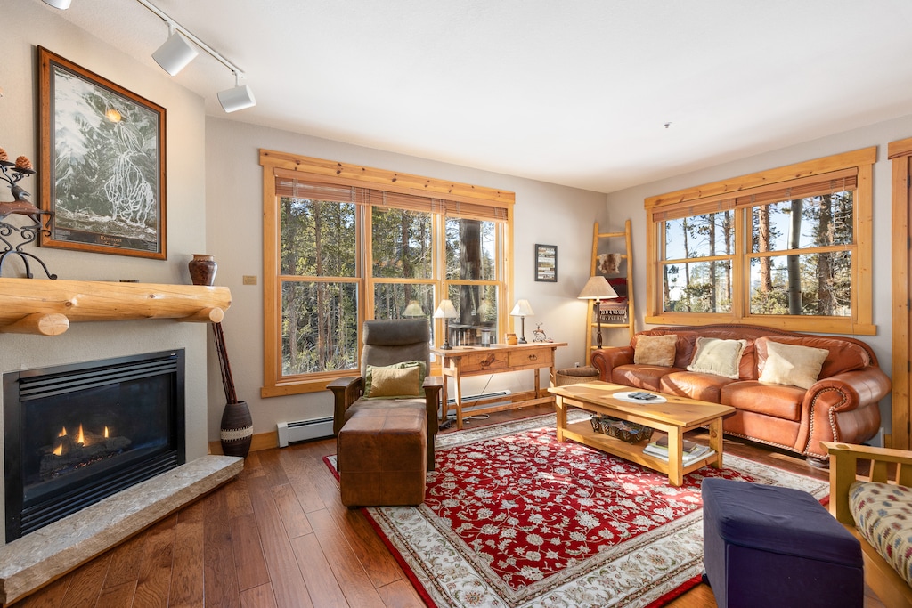 A cozy living room with a leather sofa, armchair, a wooden coffee table, a fireplace on the left, large windows with wooden frames, and a red patterned rug on a wooden floor.