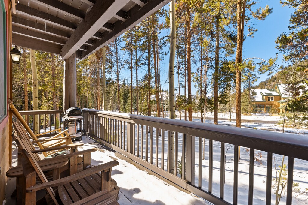 A wooden deck with rocking chairs and a grill overlooks a snowy landscape with tall trees and houses in the background.