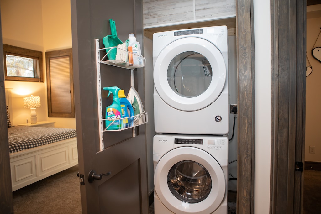 A laundry closet with a stacked washer and dryer, cleaning supplies on the door rack, and a glimpse of a bedroom on the left.