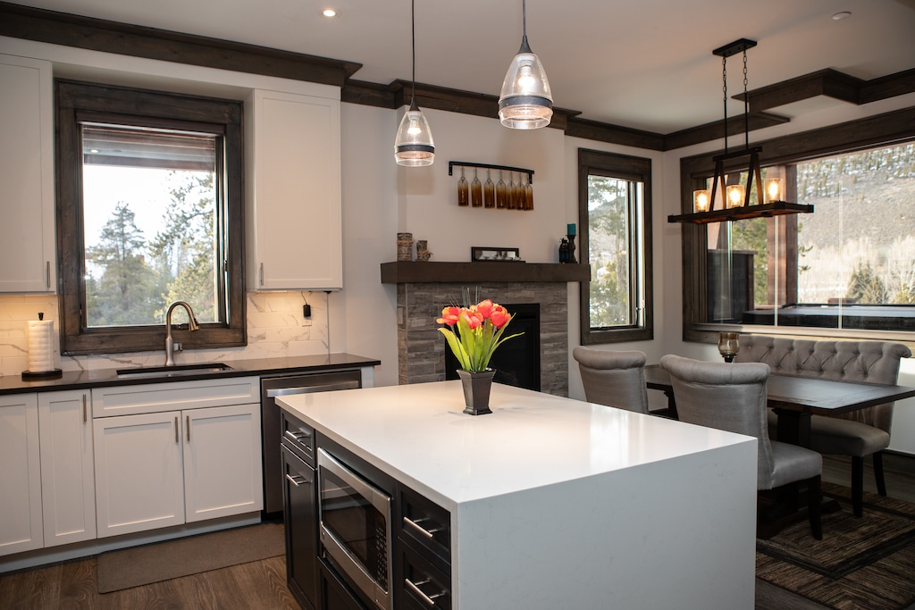 Modern kitchen and dining area with a central island, pendant lights, white cabinets, a fireplace, large windows offering outdoor views, and a vase of tulips on the island.