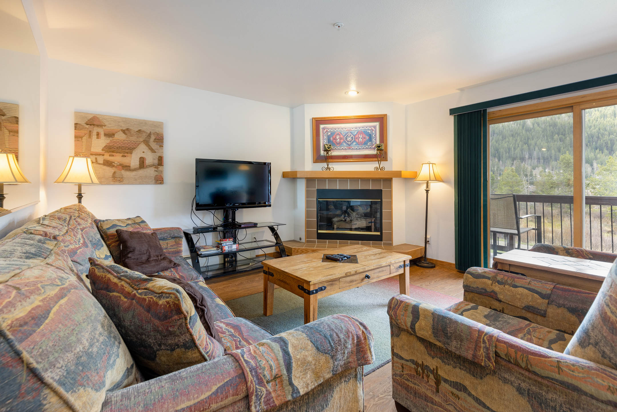 A cozy living room features patterned couches, a wooden coffee table, a TV, a fireplace, and framed art. A sliding glass door opens to a balcony with a view of trees.