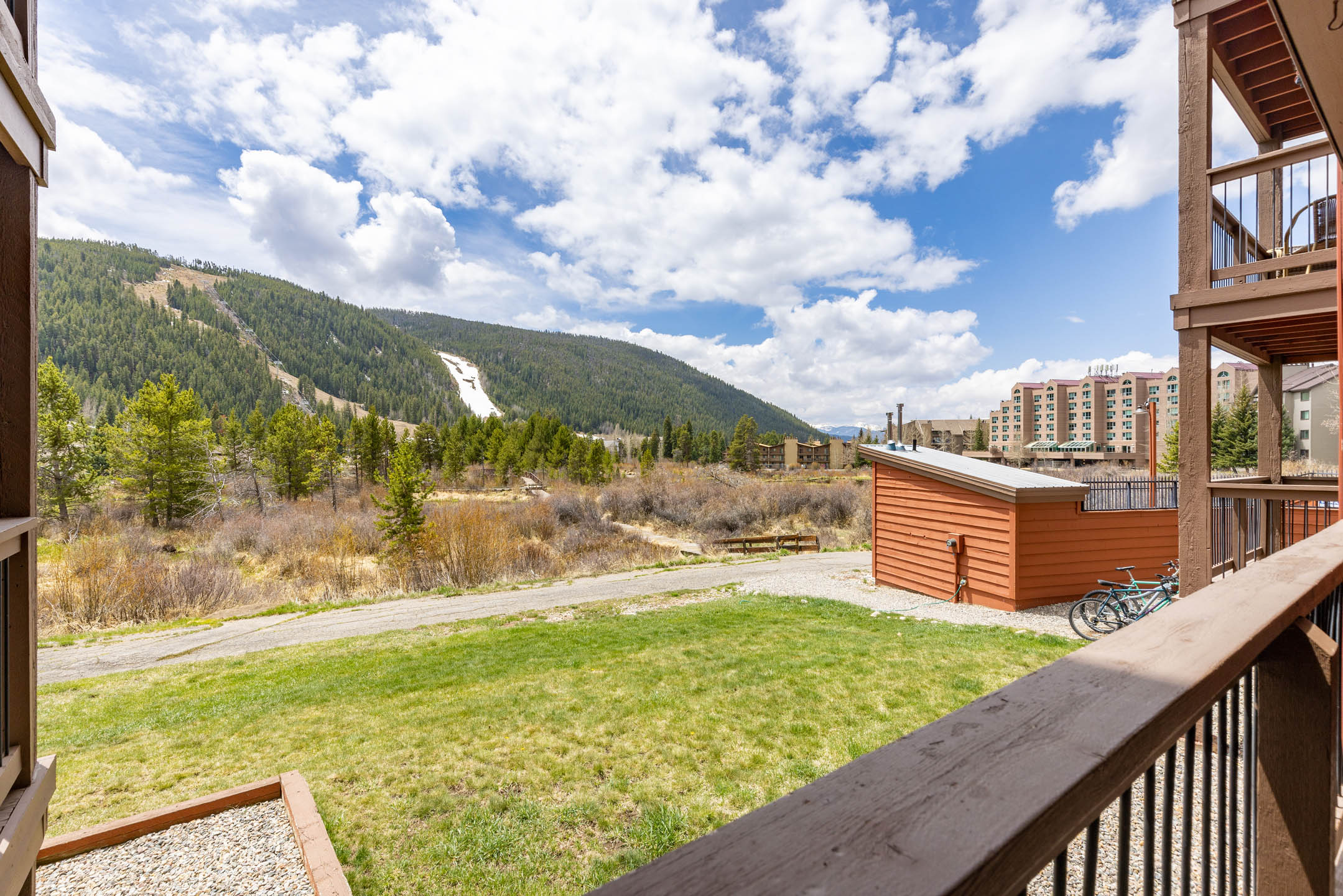 A scenic view of a mountainous landscape with a grassy lawn in the foreground, a wooden building, and several houses in the background under a partly cloudy sky.