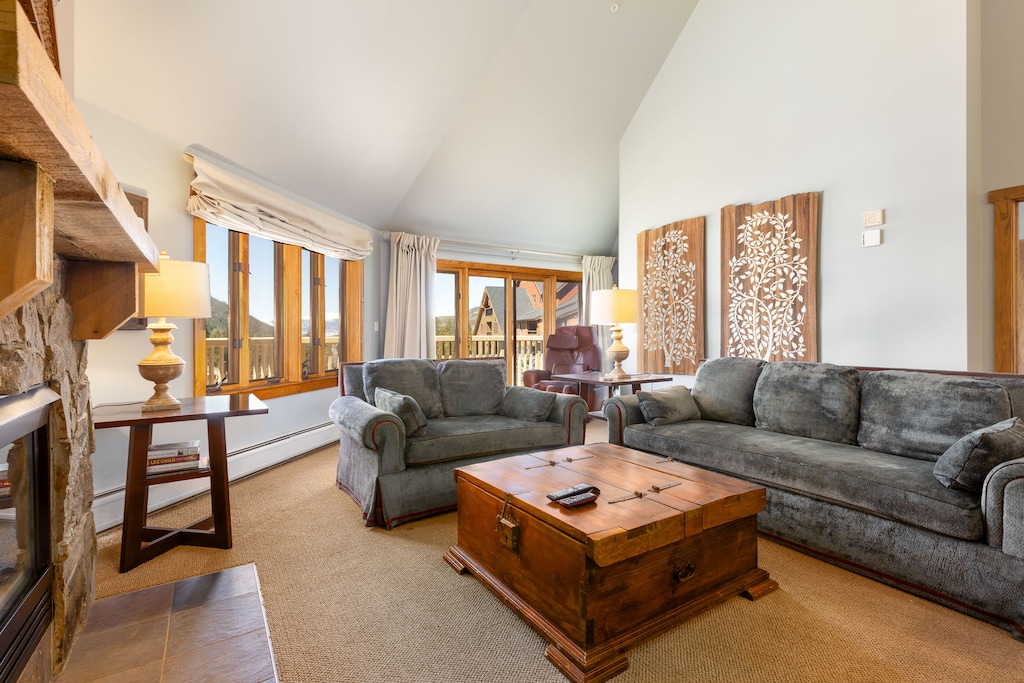 A living room with high ceilings, two sofas, a large wooden coffee table, a stone fireplace, and a window offering a view of the outdoors. The room is decorated with lamps and wall art.