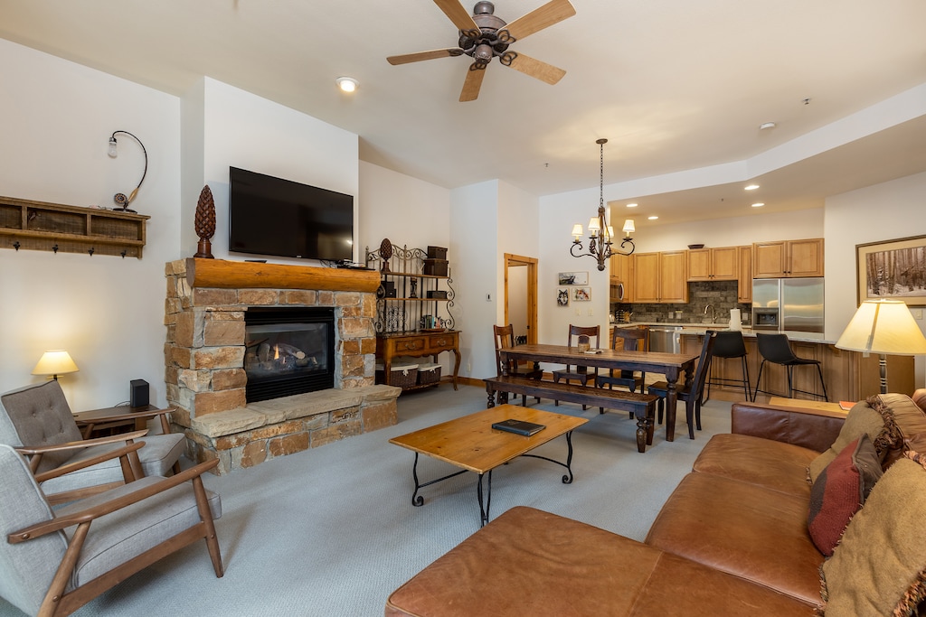 A cozy living room features a stone fireplace, a mounted TV, and a wooden coffee table. The adjacent dining area includes a long table with benches. A kitchen with stainless steel appliances is visible in the background.
