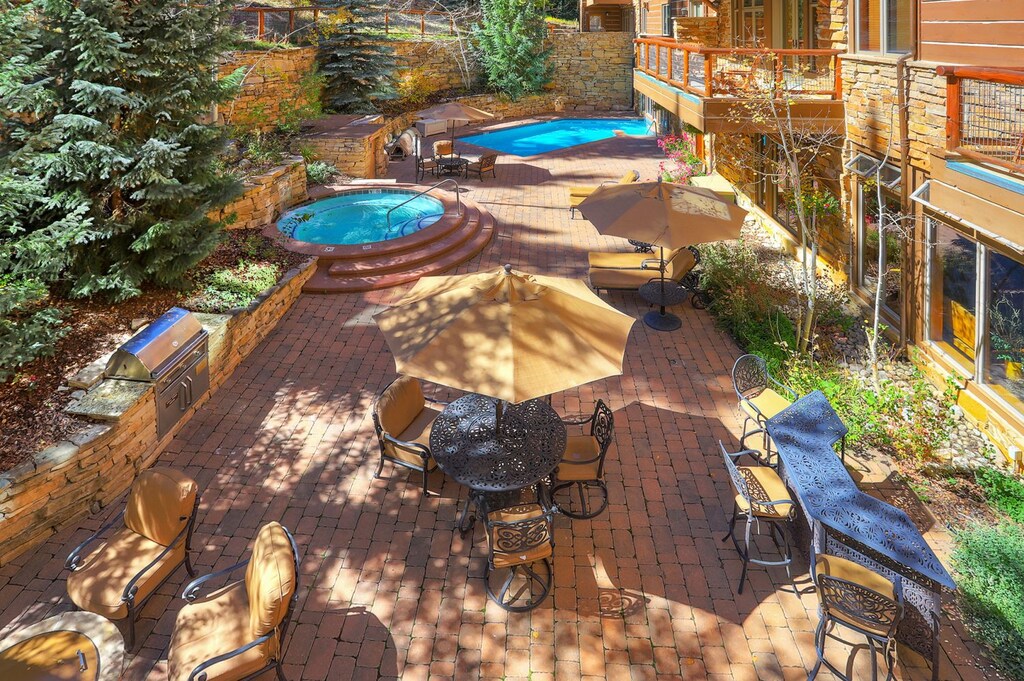 A brick patio features table sets with umbrellas, a grill, a seating area, a circular hot tub, and a rectangular swimming pool. Trees and stone walls decorate the surrounding area.