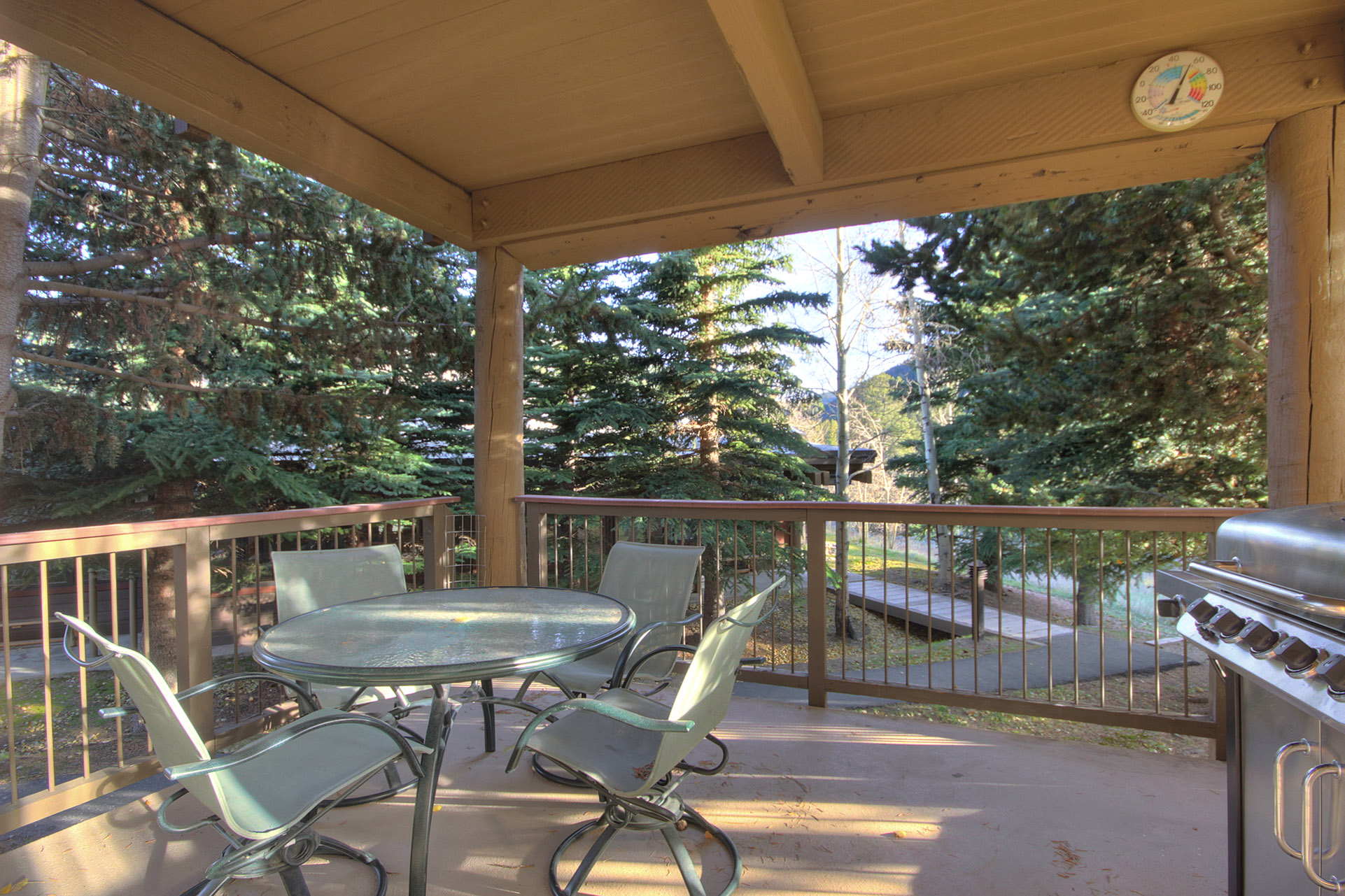 A covered outdoor patio with a metal table and four chairs, a barbecue grill, surrounded by trees and overlooking a forested area.