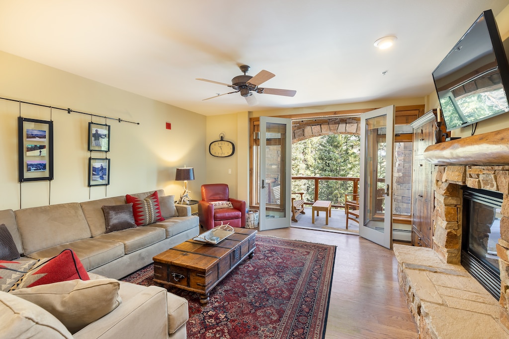 A living room with a red armchair, beige sofa, wooden coffee table, wall-mounted TV, and fireplace. Open double doors lead to a balcony with outdoor seating and view of trees.