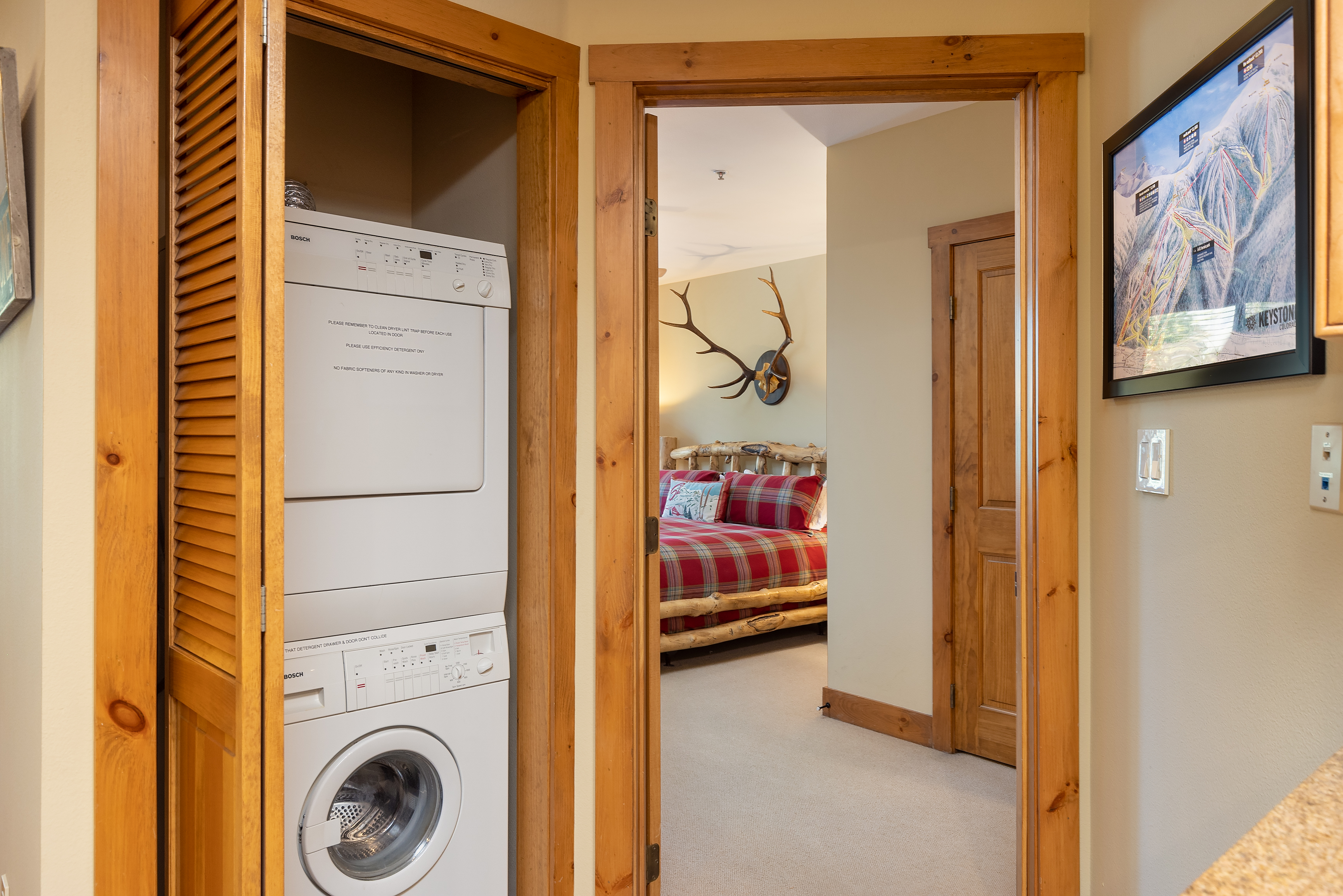 A compact laundry area with a stacked washer and dryer unit is situated next to an open doorway leading to a bedroom with a plaid bedspread and mounted deer antlers on the wall.