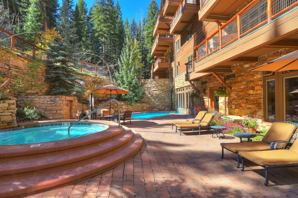 Outdoor patio with hot tub, swimming pool, lounge chairs and umbrellas, bordered by tall trees and a multi-story stone building on a sunny day.