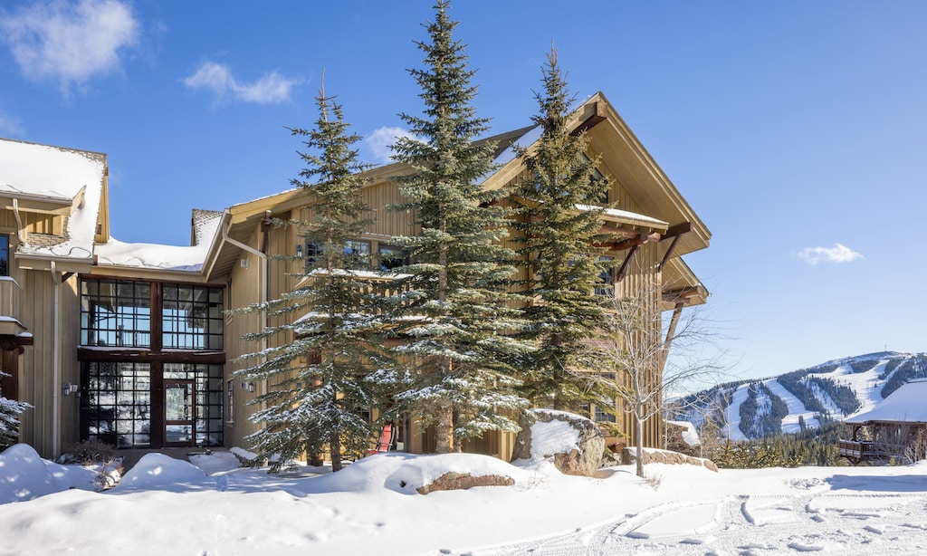 Mountain lodge surrounded by snow-covered evergreens with ski slopes visible in the distance under clear winter skies.