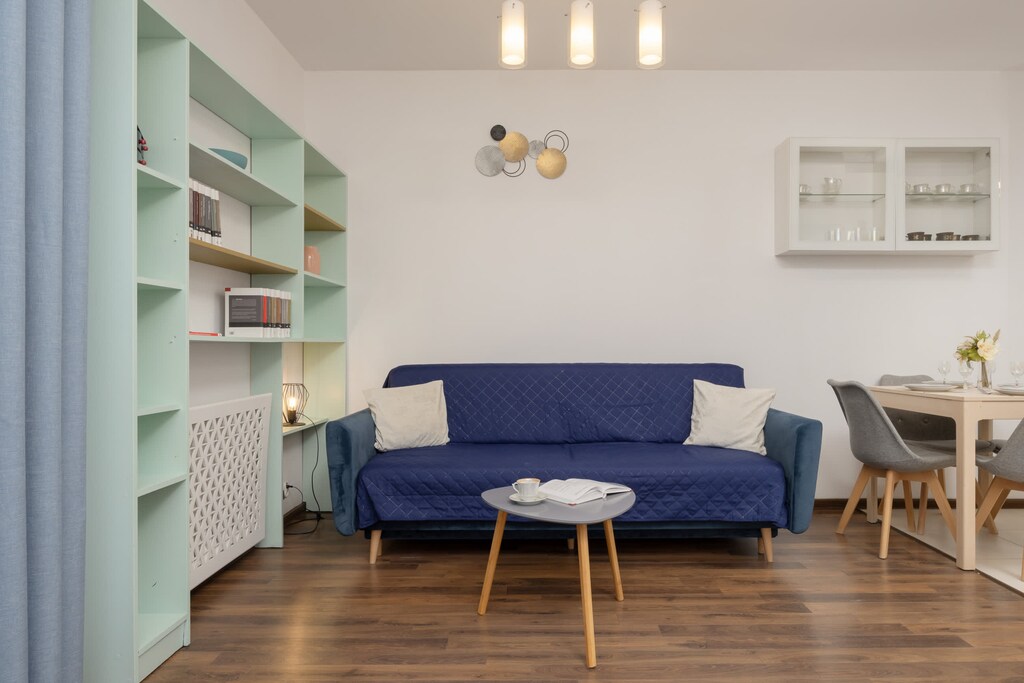 A living room with a blue sofa and light wood flooring. The sofa is complemented by a small round coffee table, and the wall behind it features built-in shelving.
