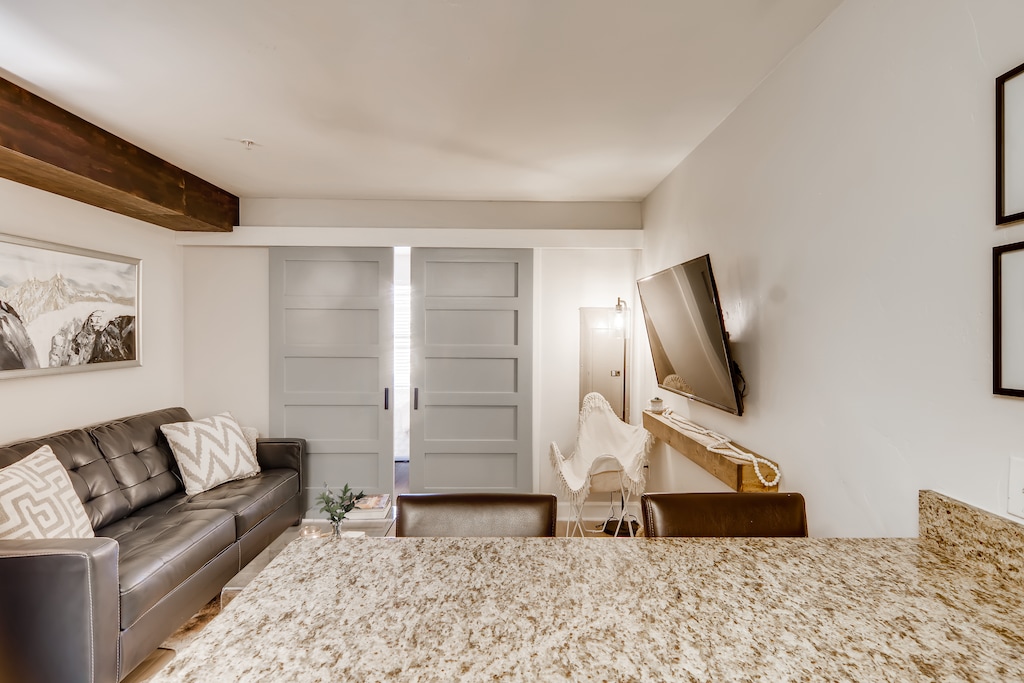 A modern living room with a granite countertop in the foreground, a black leather sofa, a wall-mounted TV, and light gray sliding doors in the background. Light fixture and framed pictures are also visible.