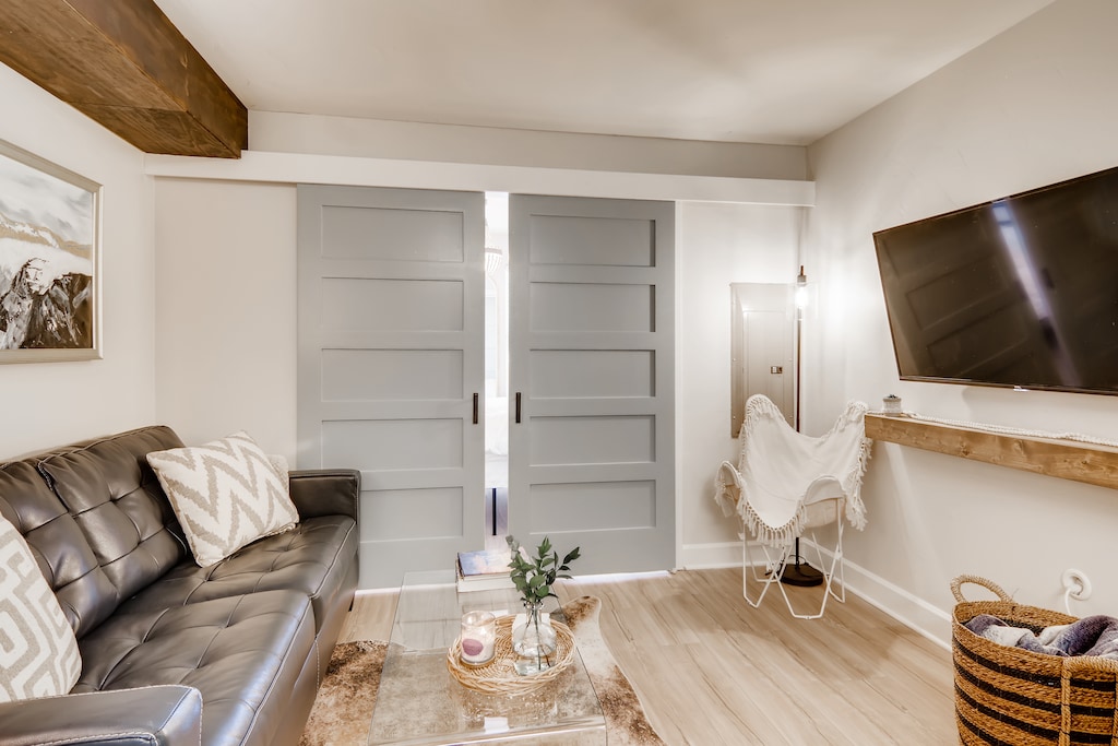 A contemporary living room with a black leather sofa, wooden coffee table, TV mounted on the wall, and gray sliding doors. A small chair and a basket are also present.