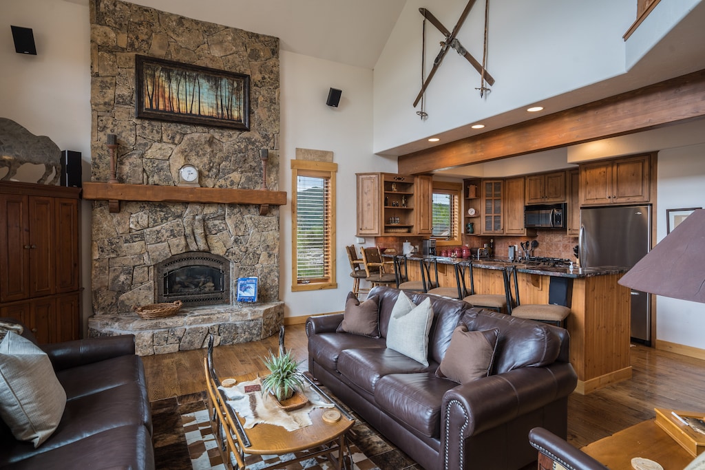 A cozy living room features a stone fireplace with a wooden mantel, leather sofas, a rustic coffee table, and a kitchen area with wooden cabinets and a breakfast bar in the background.