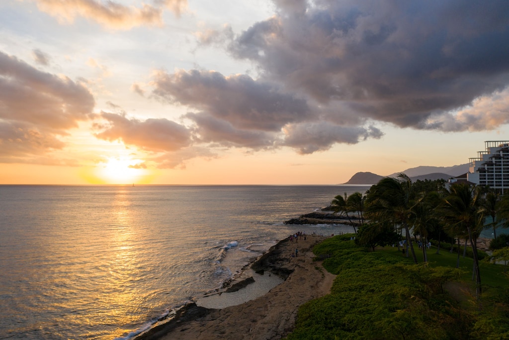 Stunning beachfront location with golden sunset views over the ocean, palm trees, and nearby resort buildings creating a tropical paradise setting.