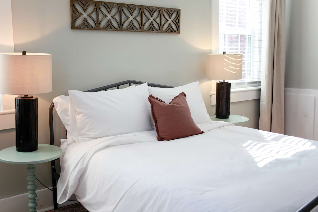 Bedroom with a queen size bed and nightstands with lamps and artwork. Windows flanking each side of the bed. 