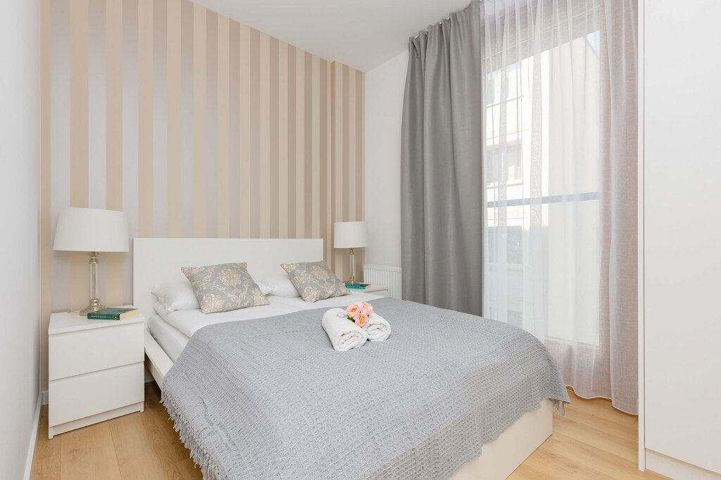 A stylish bedroom with a double bed, light gray bedding, and striped wallpaper in a soft, neutral color.
