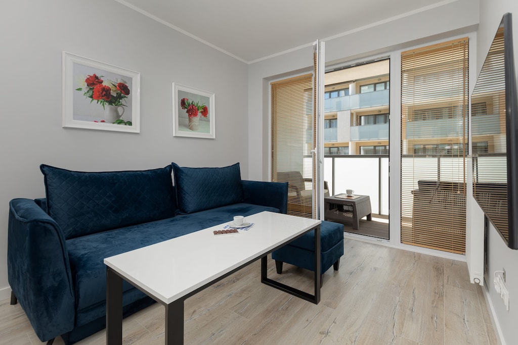 A cozy living room with a dark blue L-shaped couch, a coffee table, and a flat-screen TV mounted on the wall. Large windows allow plenty of natural light.