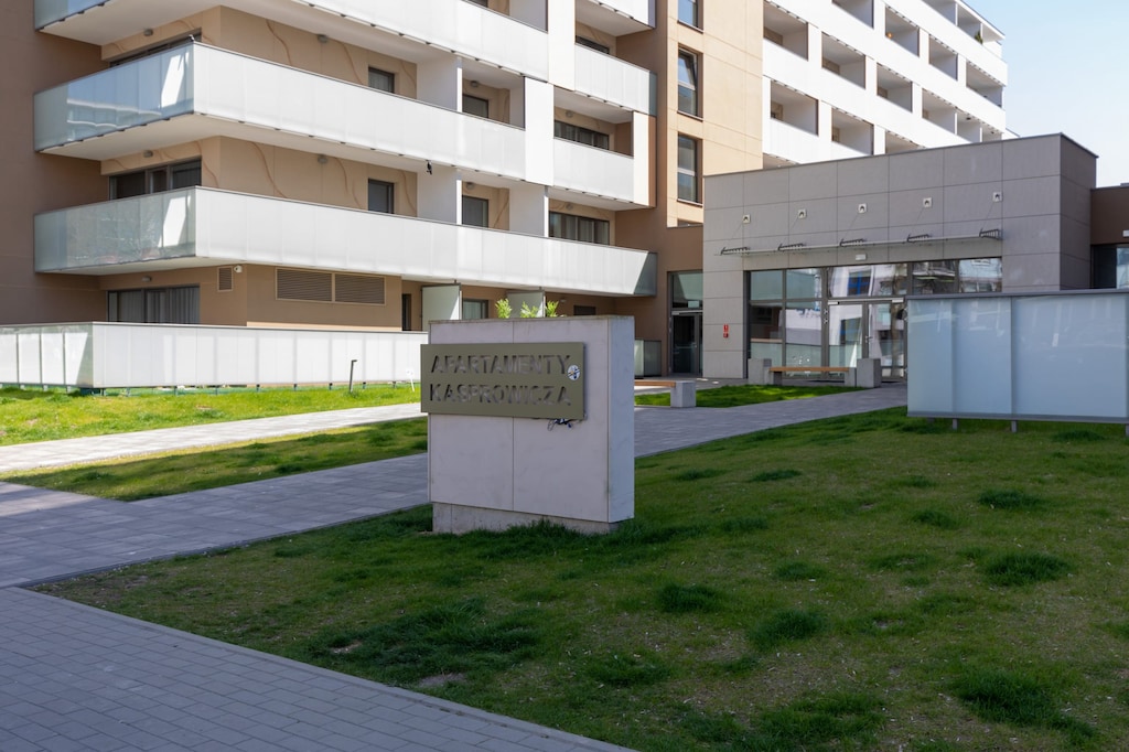 The exterior view of the apartment complex shows modern buildings, green grass, and a paved walkway.

