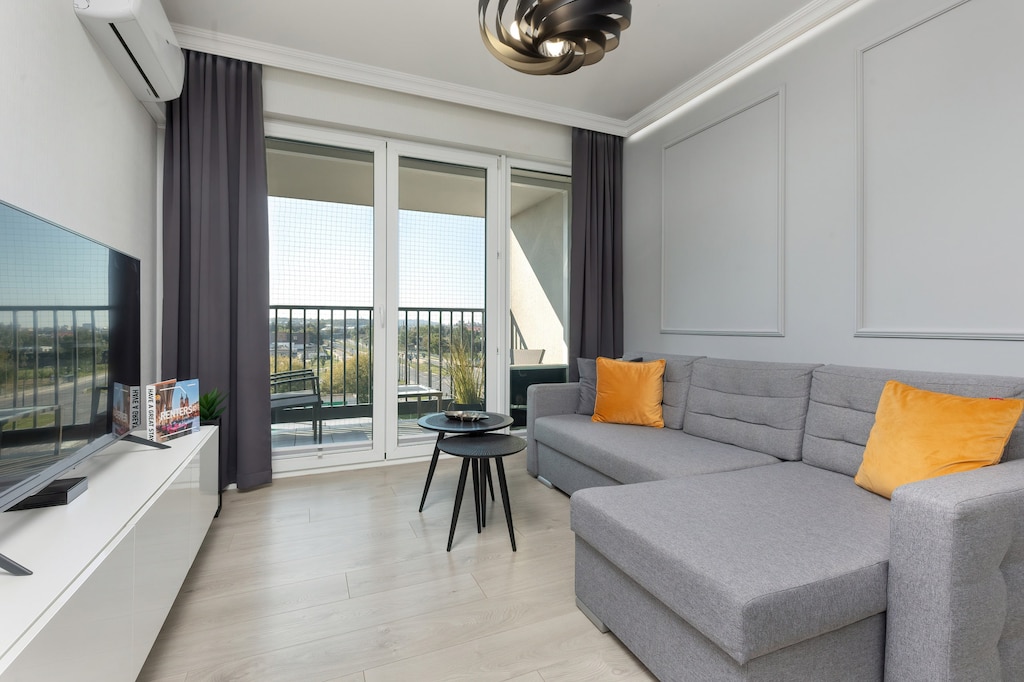 A cozy living room with a gray sofa, yellow accent pillows, and a coffee table, complemented by natural light streaming through large windows.
