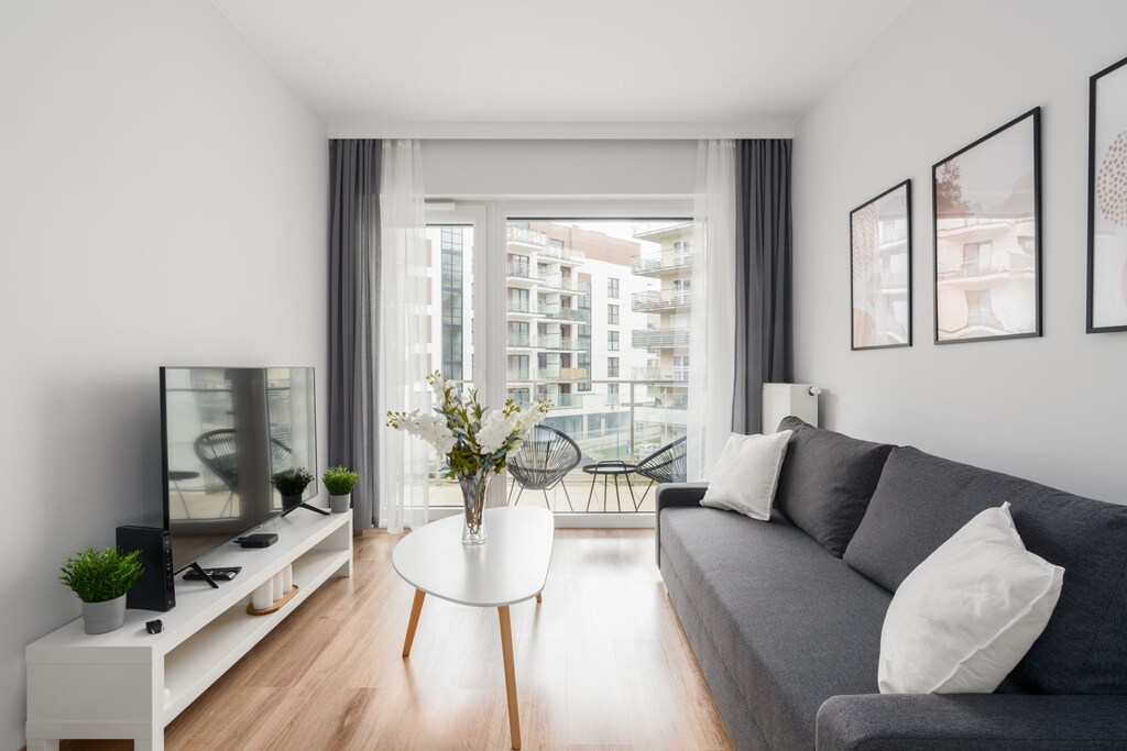 A cozy living room with a gray sofa, a coffee table, and a TV, complemented by a city view from large windows.
