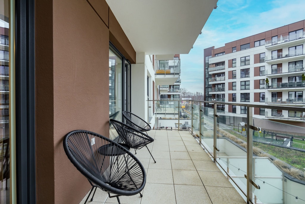 A spacious balcony with a glass railing and a modern chair, overlooking a contemporary apartment complex.
