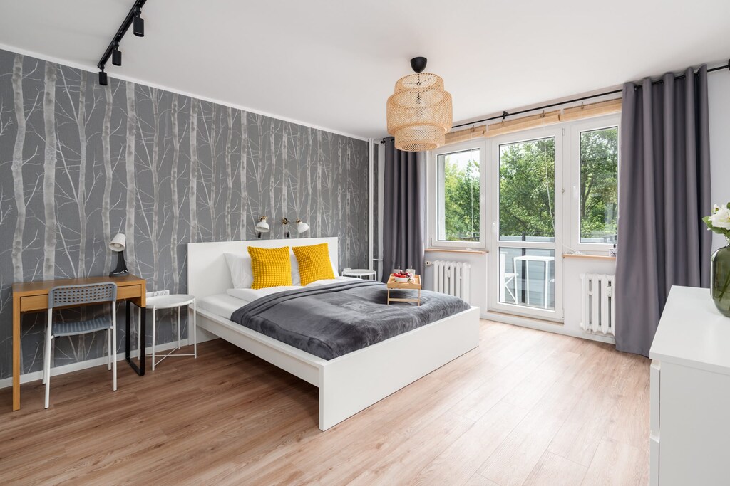 A stylish bedroom with a large bed, gray and yellow accents, and a spacious window overlooking greenery.


