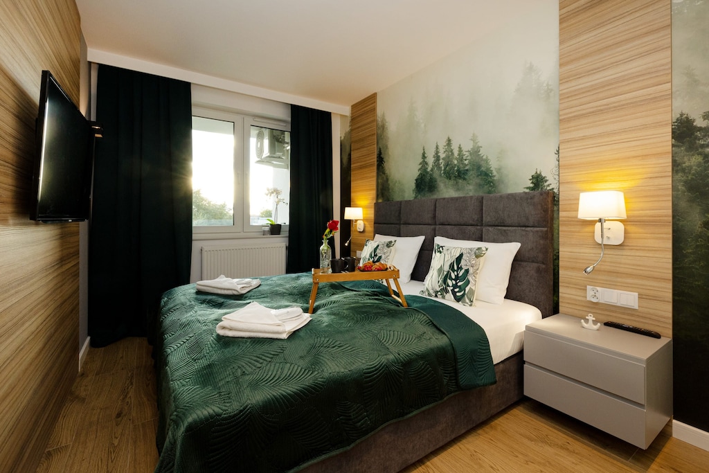A cosy bedroom combining the naturalness of wood with a wallpaper depicting a misty forest. A flat-screen TV hangs on the wall and dark curtains help to darken the room.