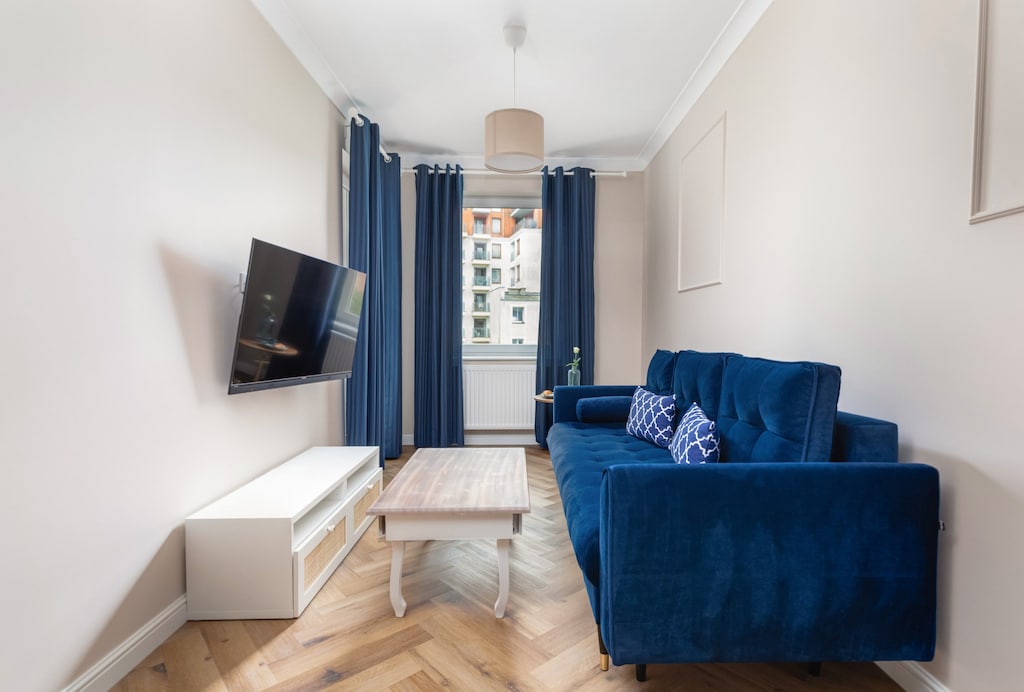 A cozy living room featuring a deep blue sofa, a wall-mounted TV, and natural light from large windows.

