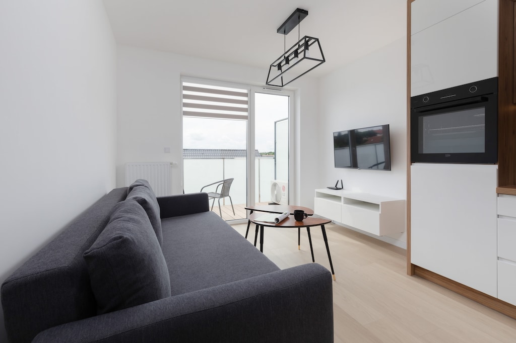 A modern living room featuring a gray sofa, wooden coffee table, and a wall-mounted TV, with bright natural lighting from large windows.