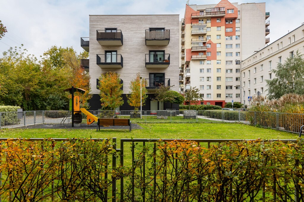 A view of a green park with bushes and trees, surrounded by modern apartment buildings.