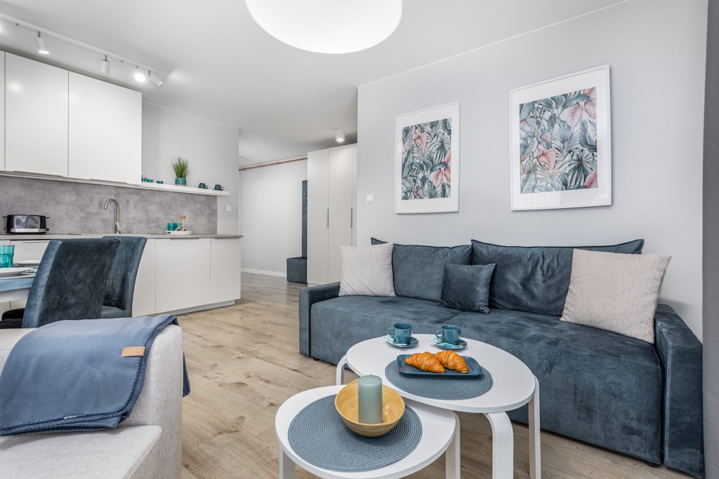 A modern yet cosy space. with a grey soft sofa over which two paintings hang. In the background you can see the kitchenette with white cabinets.