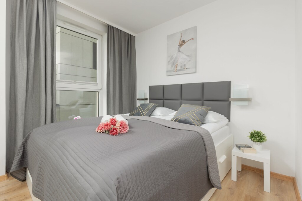 A cozy bedroom featuring a grey bedspread, a white nightstand, and sheer curtains leading to the balcony.
