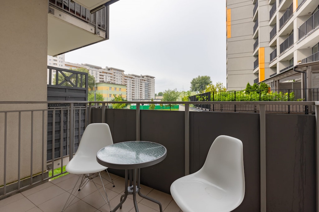 A small outdoor balcony with a round table and two chairs, overlooking a modern urban setting.
