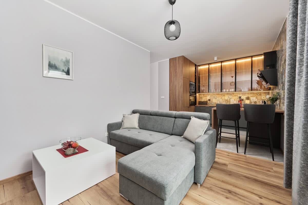 A bright and spacious living area with a large gray sofa, coffee table, and a TV on a sleek white wall unit. The room has natural light streaming in through big windows.