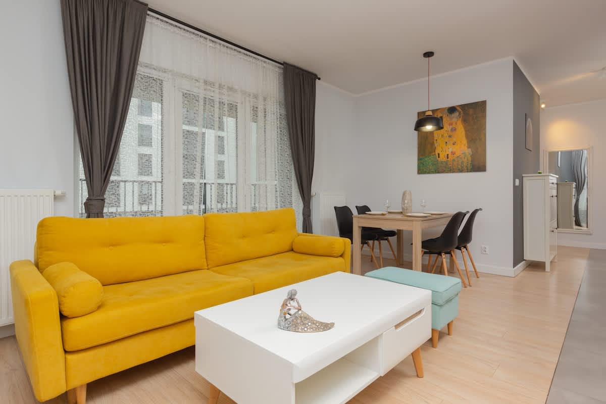 A modern living room featuring a bright yellow sofa, a white coffee table, and a TV on a stand. Large windows with sheer and blackout curtains bring in natural light.