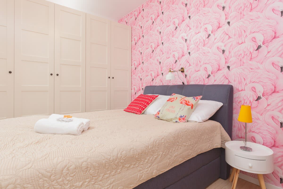 A cozy bedroom with a queen-sized bed against a wall decorated with pink floral wallpaper. The room includes bedside tables and a lamp.