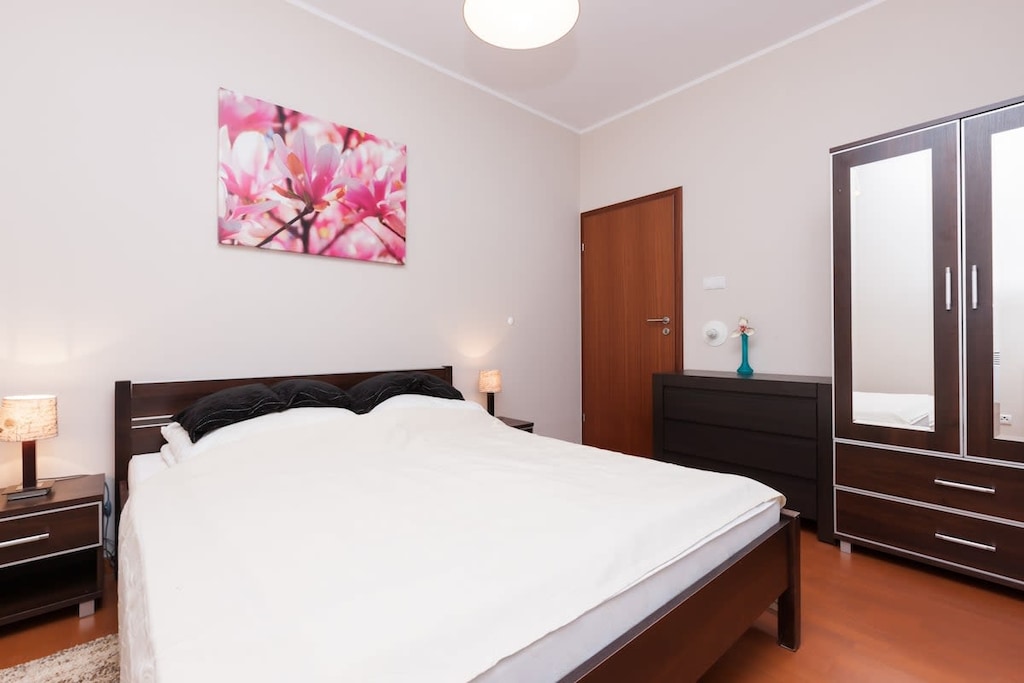 A bright bedroom with a double bed, a pink floral painting, and wooden furniture.
