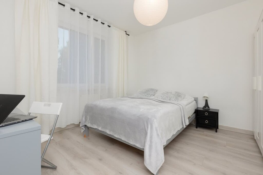 A cozy bedroom featuring a double bed with white linens, a bedside table, and sheer curtains allowing light to come through.
