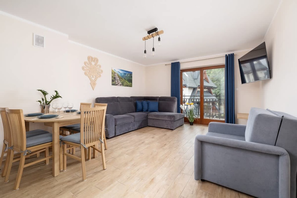 A cozy living room with a wooden dining table, chairs, and a sofa. Large windows let in natural light and offer a bright atmosphere.