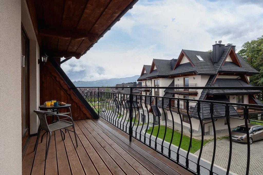 A wooden-floored balcony with a metal railing, overlooking a peaceful neighborhood with scenic mountain views.