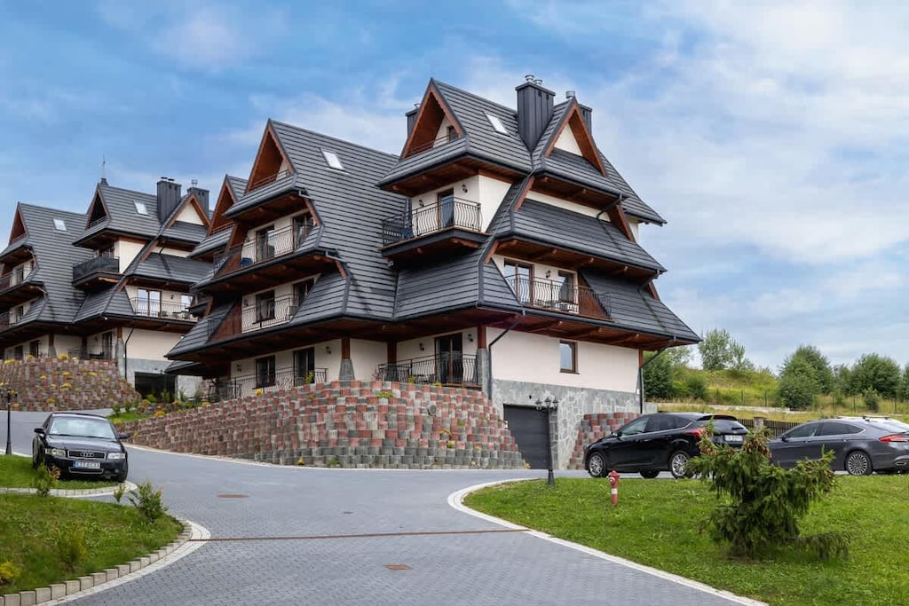 A chalet-style building with a wooden roof and stone foundation surrounded by green grass and a paved driveway.