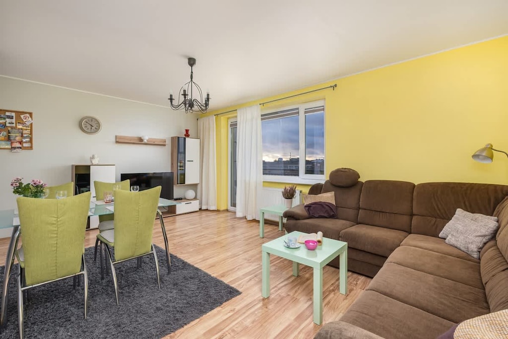 A bright living-dining area with yellow walls, a brown corner sofa, and a dining table with green chairs, creating a cozy and cheerful atmosphere.
