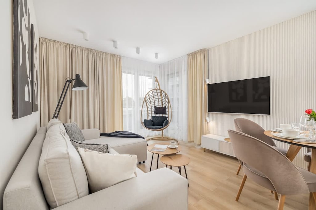A bright living room with a beige sofa, a TV mounted on the wall, and a hanging chair near the window.
