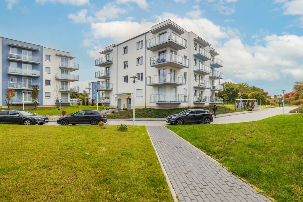 A modern residential building with white facades, surrounded by green lawns and a walking path.
