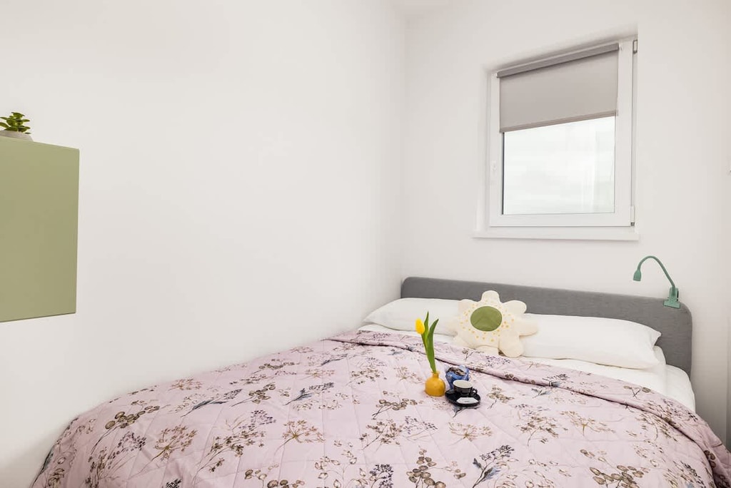 A cozy bedroom with a floral bedspread, a small window, and a green bedside shelf.

